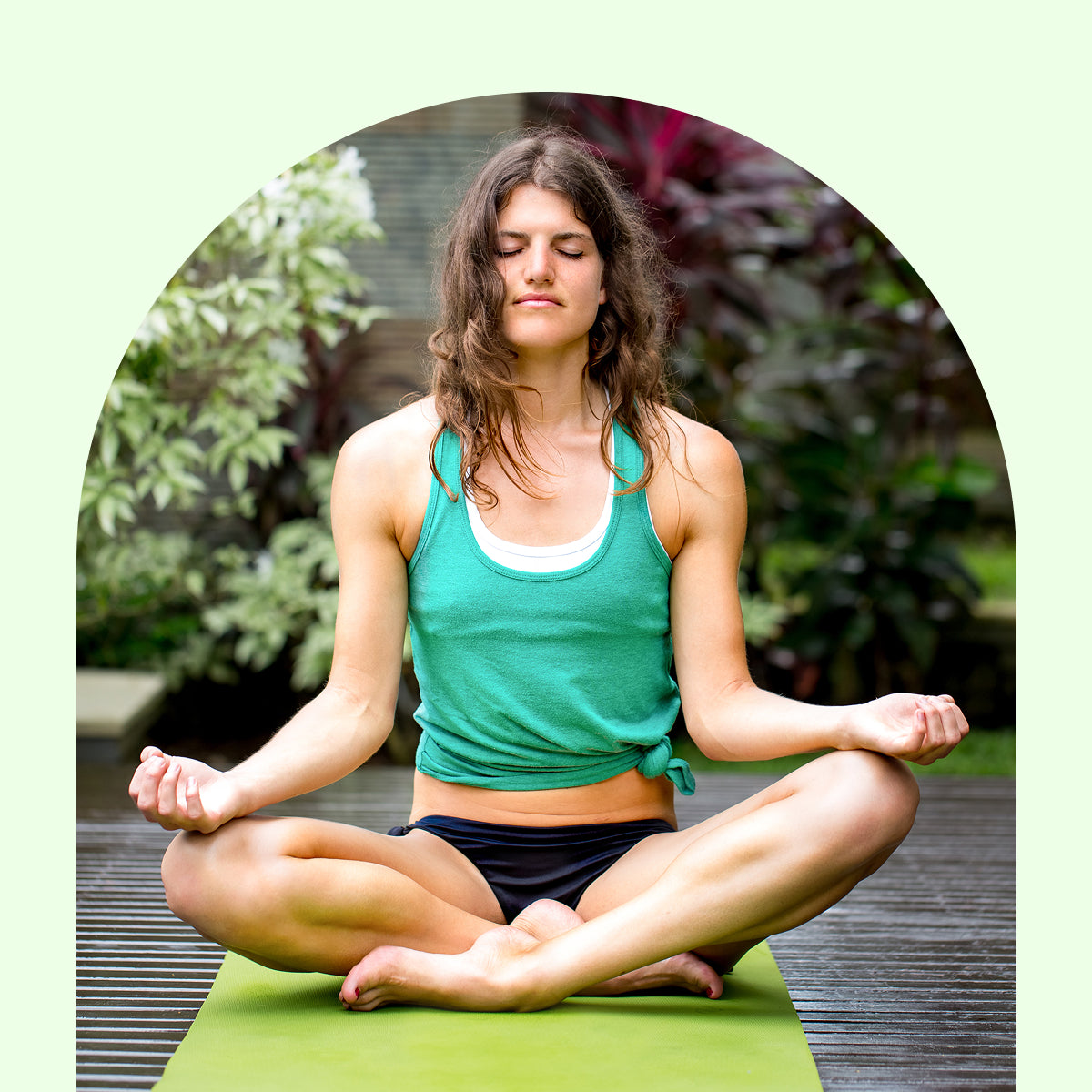 Woman in green tank top and black shorts meditating on a green mat outdoors.